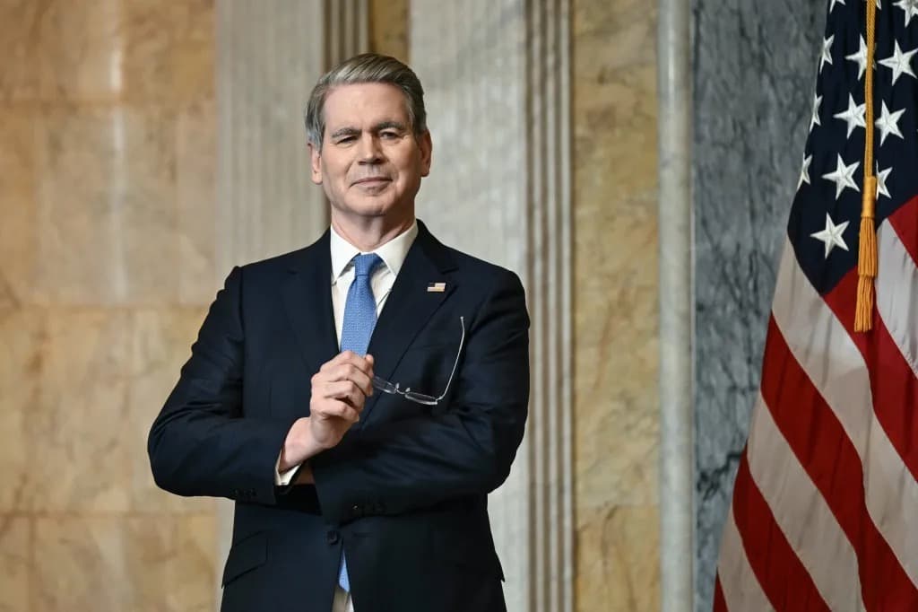 Treasury Secretary Scott Bessent standing in a marble hallway holding his glasses, next to a US flag