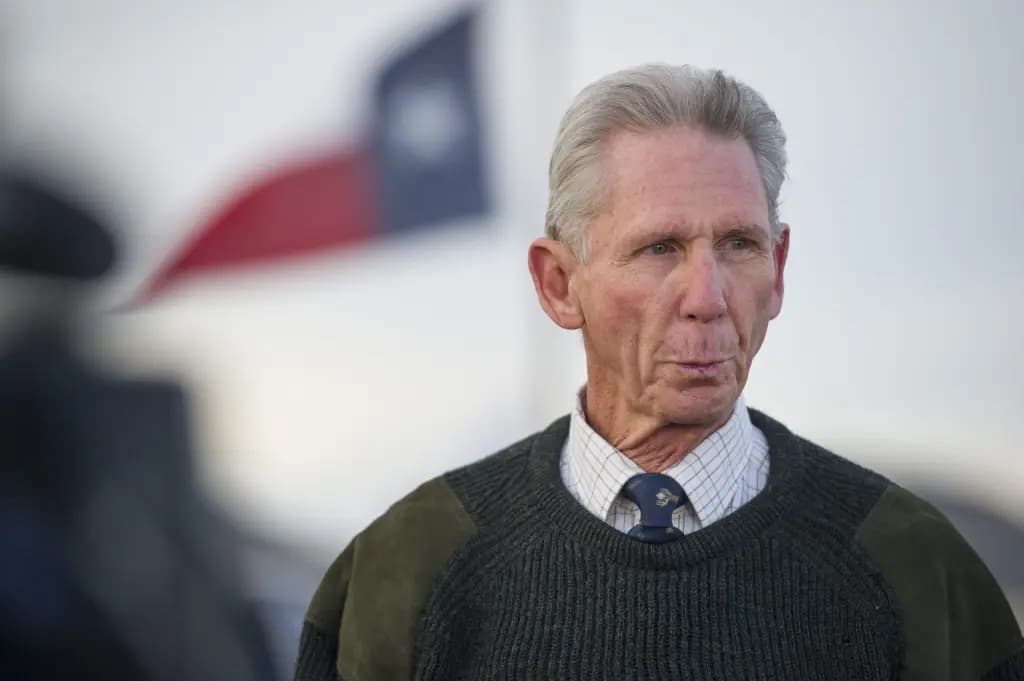 An older man standing outdoors in front of a Texas flag