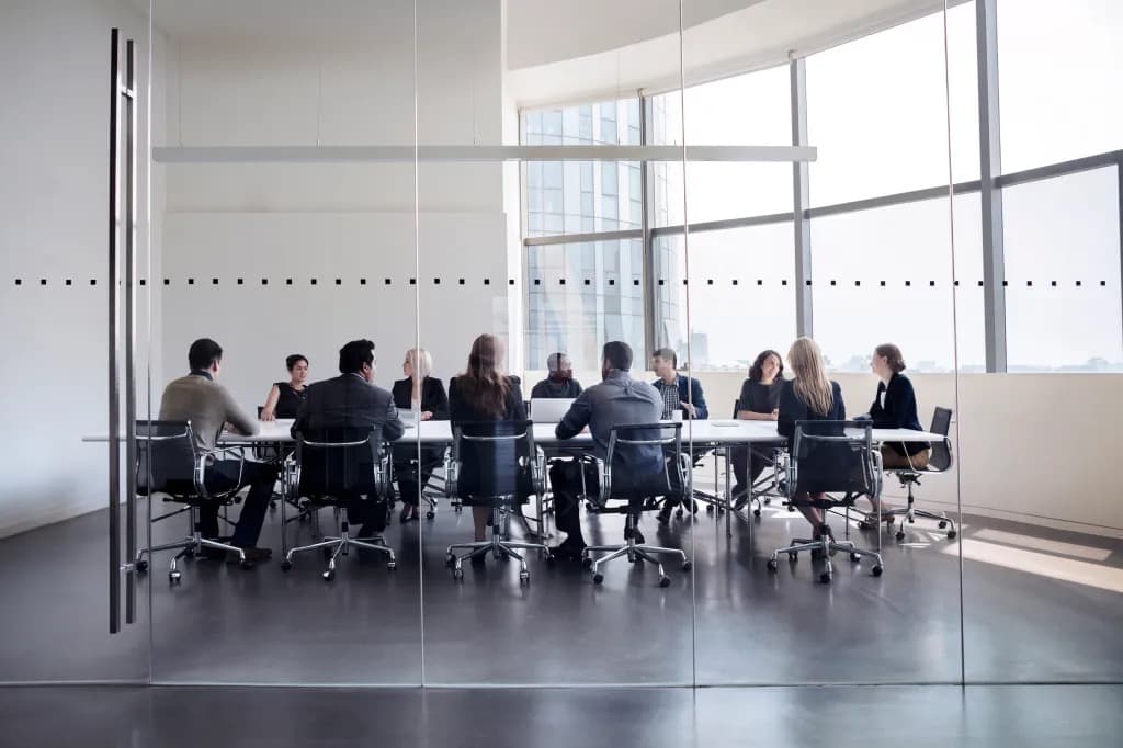 A group of colleagues seated around a conference table in a glass walled meeting room