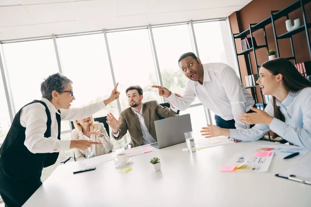 A group of colleagues arguing and pointing fingers at each other around a conference table