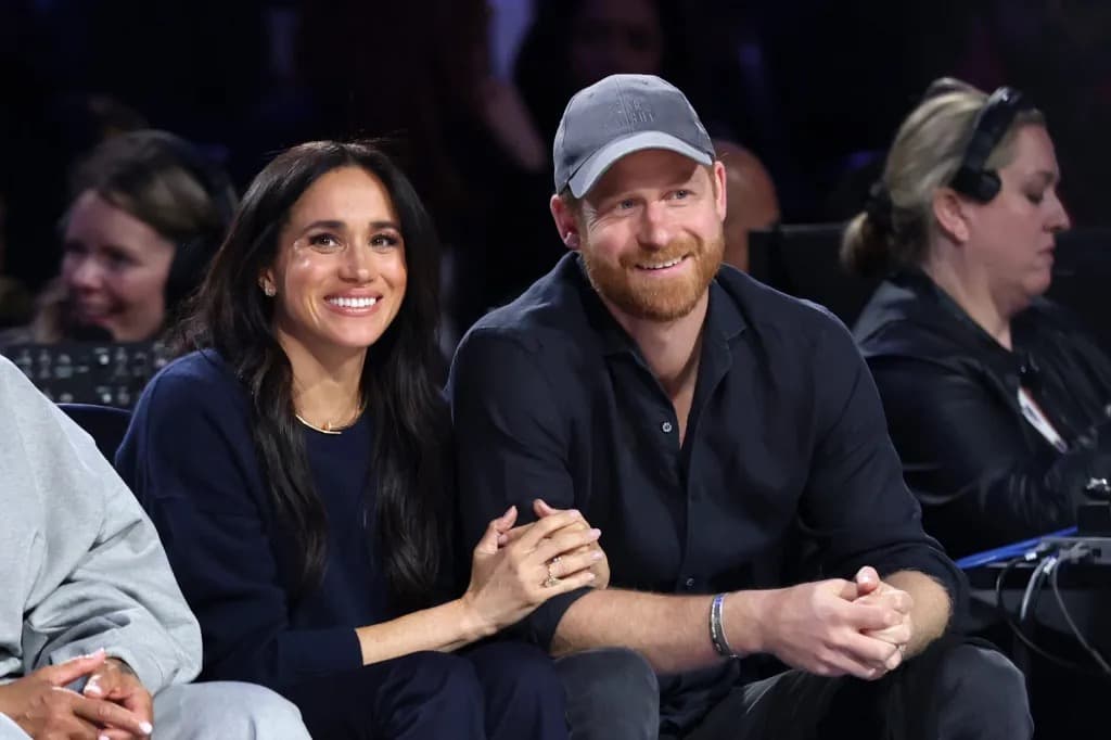 Prince Harry and Meghan smiling and holding hands while seated at an event
